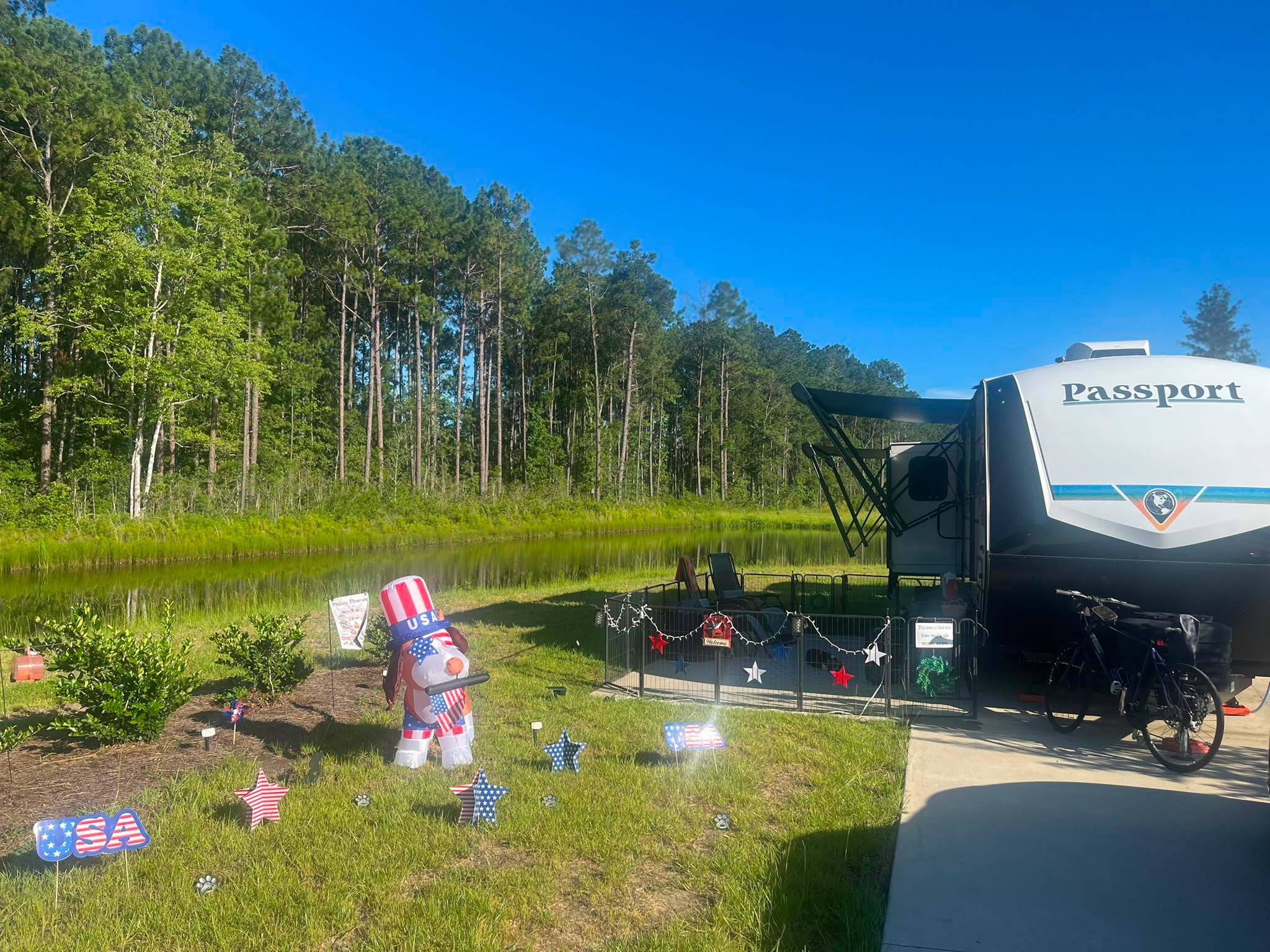 A camper labeled "Passport" is parked near a pond with a bicycle, patriotic USA decorations, and trees in the background under a clear blue sky.