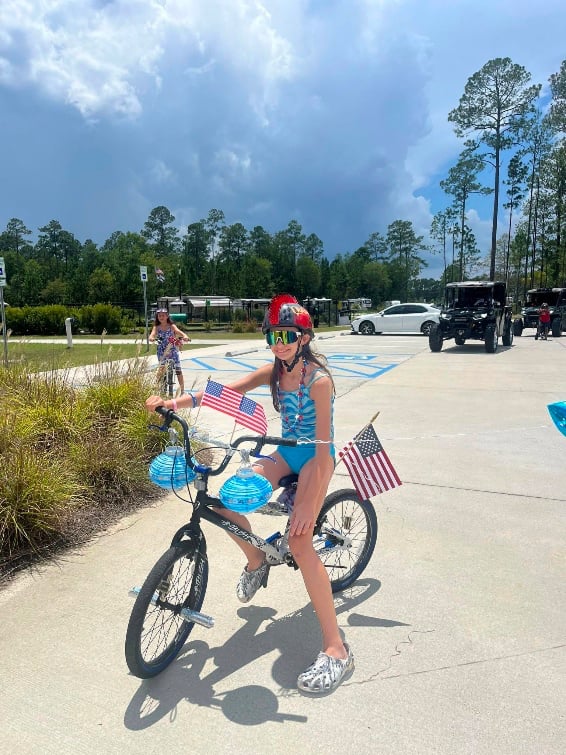 A girl wearing a helmet and sunglasses sits on a decorated bicycle with American flags in a parking lot on a sunny day.
