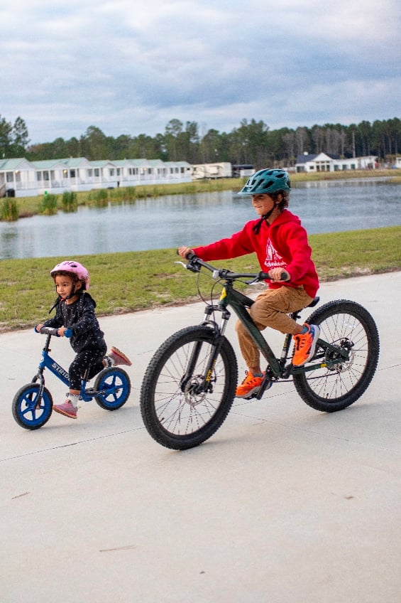 A young girl on a balance bike and an older boy on a bicycle ride along a paved path near a lake, both wearing helmets.