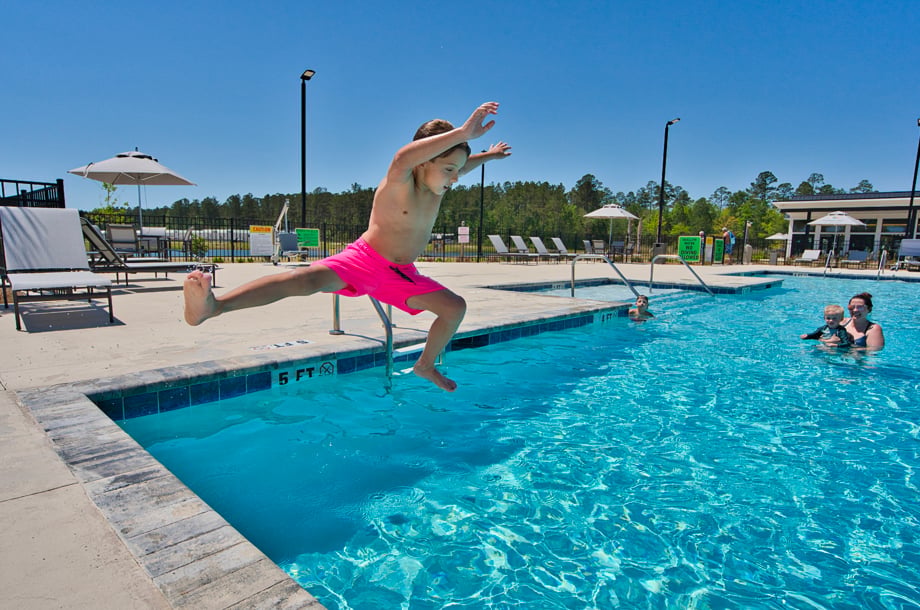 A young boy in pink swim trunks jumps into an outdoor pool while others watch and swim on a sunny day.