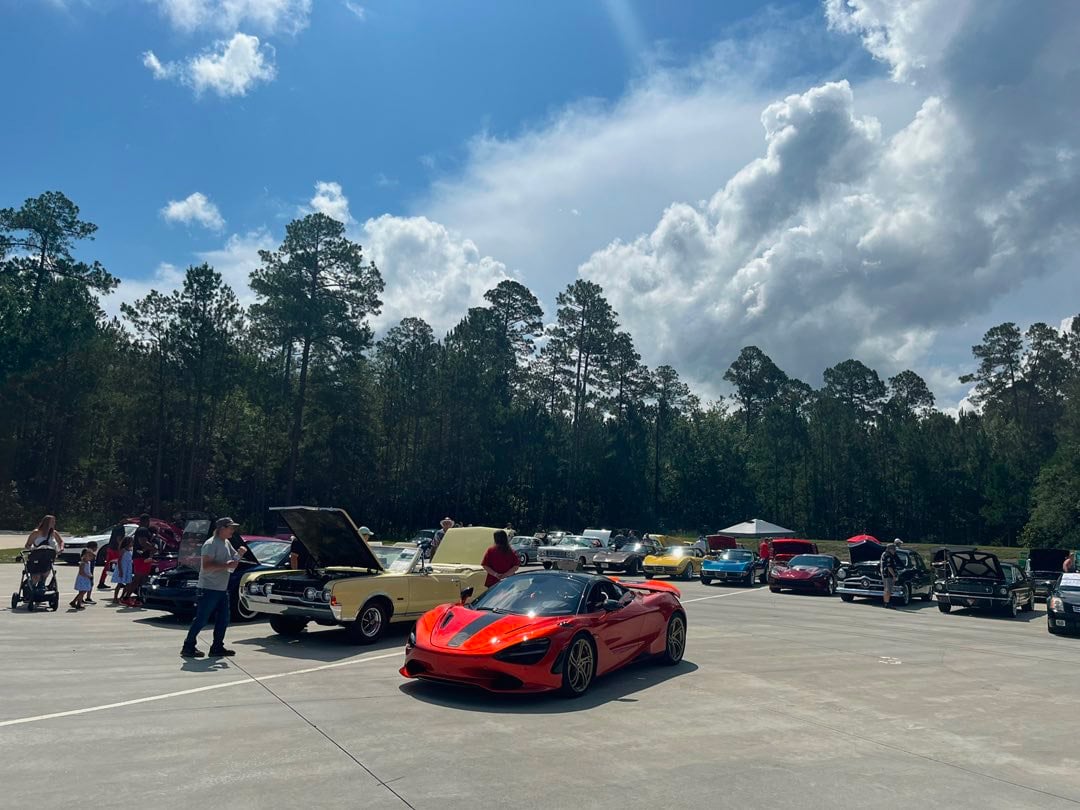 A red sports car is parked in front of several classic cars with open hoods at an outdoor car show under a partly cloudy sky.
