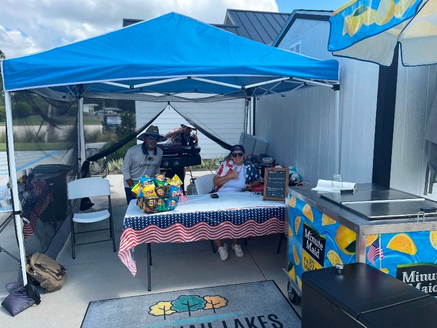 Two people sit under a blue canopy at a table decorated with an American flag and snacks, with a cooler and a Minute Maid stand nearby.