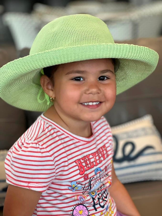 Young child wearing a large light green sunhat and a striped shirt sits on a couch, smiling at the camera. Pillows are visible in the blurred background.