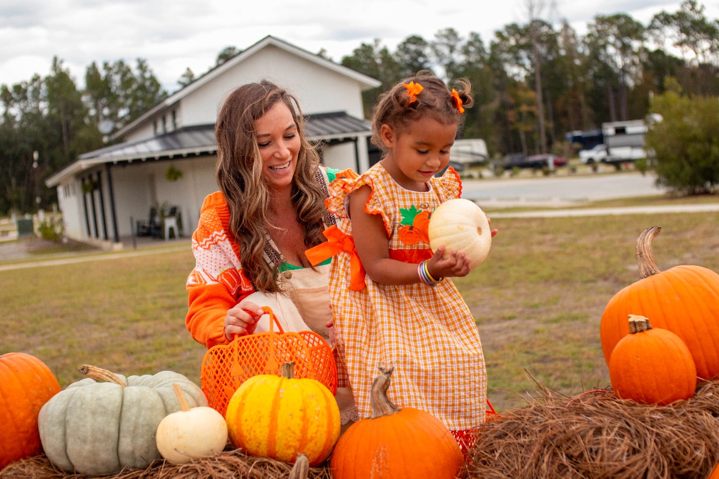 An adult and a child in orange clothing look at pumpkins on a hay bale outside, with a house and trees in the background.