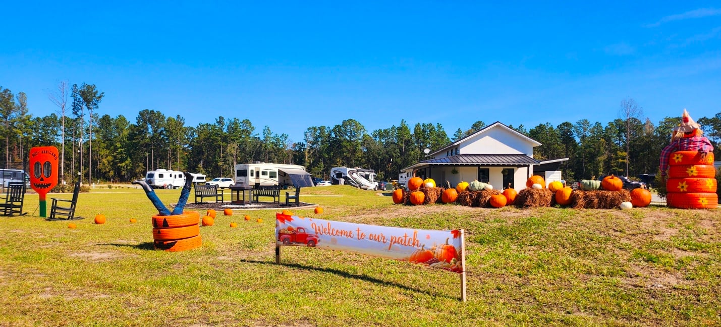 A pumpkin patch with orange pumpkins, hay bales, a welcome banner, a small building, and RVs in the background on a sunny day.