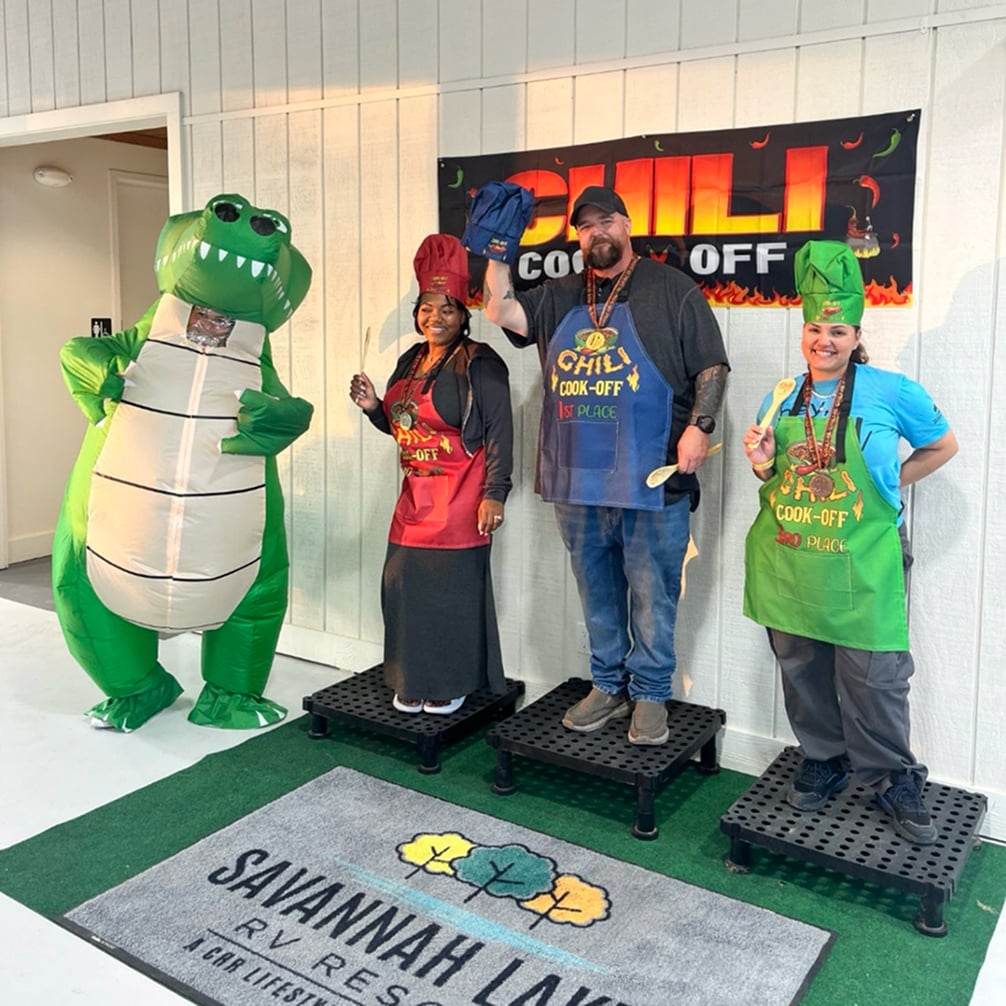 Three people in aprons and chef hats stand on podiums with medals at a chili cook-off event, with a person in an inflatable alligator costume beside them. A banner and event signage are visible.