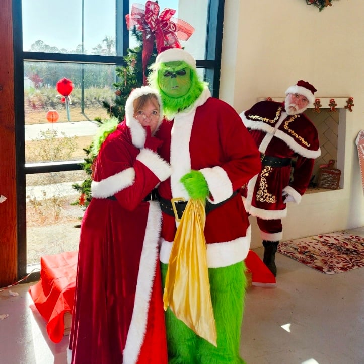 Three people in holiday costumes pose indoors: one dressed as Mrs. Claus, one as the Grinch in a Santa suit, and one as Santa Claus standing near a decorated Christmas tree.