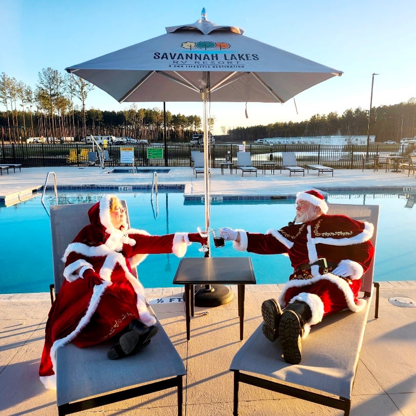 Two people dressed as Santa Claus and Mrs. Claus sit on lounge chairs by a pool, reaching towards each other under a "Savannah Lakes" umbrella.