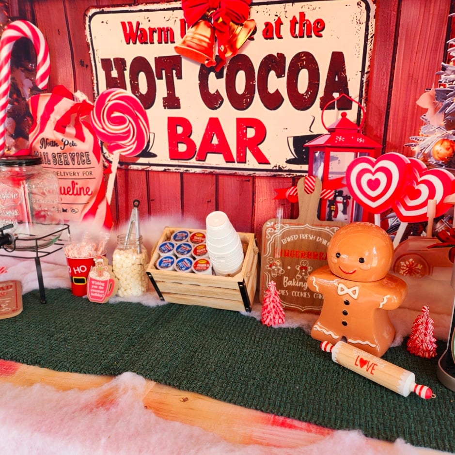 A festive hot cocoa bar with cups, cocoa pods, marshmallows, holiday decorations, a gingerbread figure, and a large “Hot Cocoa Bar” sign in the background.