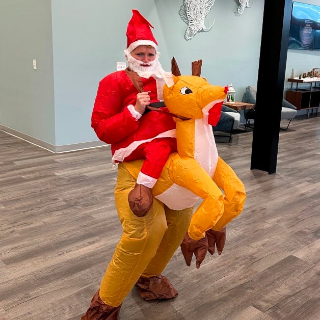 Person in an inflatable Santa riding a reindeer costume standing indoors on a wooden floor with a grayish-blue wall in the background.