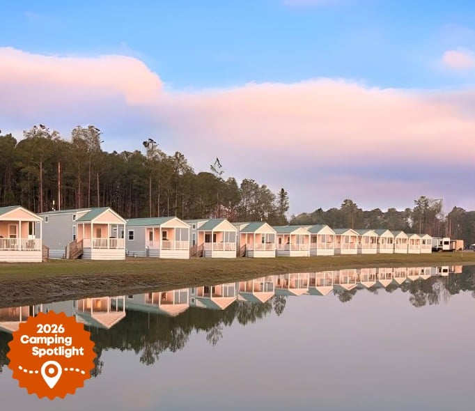 Row of small, identical cabins with porches reflected in a calm lake, bordered by trees; "2026 Camping Spotlight" badge in lower left corner.