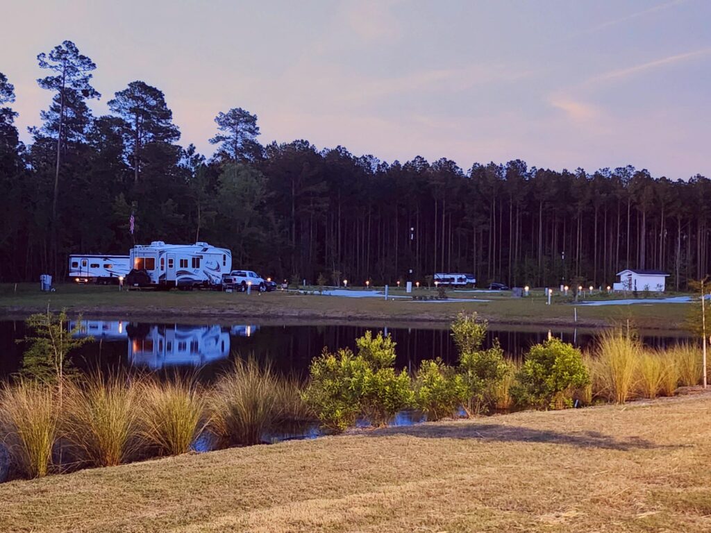 RVs and trailers are parked beside a pond with tall trees in the background at dusk. The scene reflects in the water, and short grasses are in the foreground.