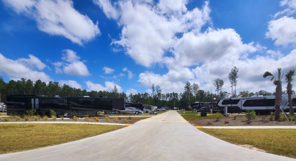 A paved road runs through an RV park with multiple large black and white RVs parked on either side, surrounded by grass, trees, and a partly cloudy sky.