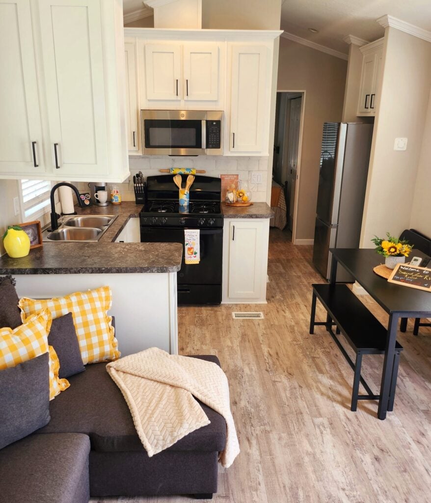 Modern kitchen and dining area with white cabinets, black appliances, gray sofa with yellow pillows, small dining table, and wood-style flooring. Sunflowers used as decor.