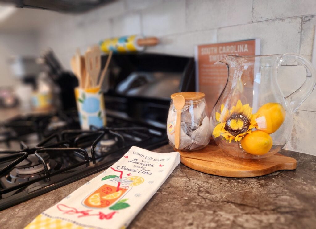 A kitchen countertop with a glass pitcher containing lemons and a sunflower, a jar of tea bags, wooden utensils, and a tea towel beside a stovetop.