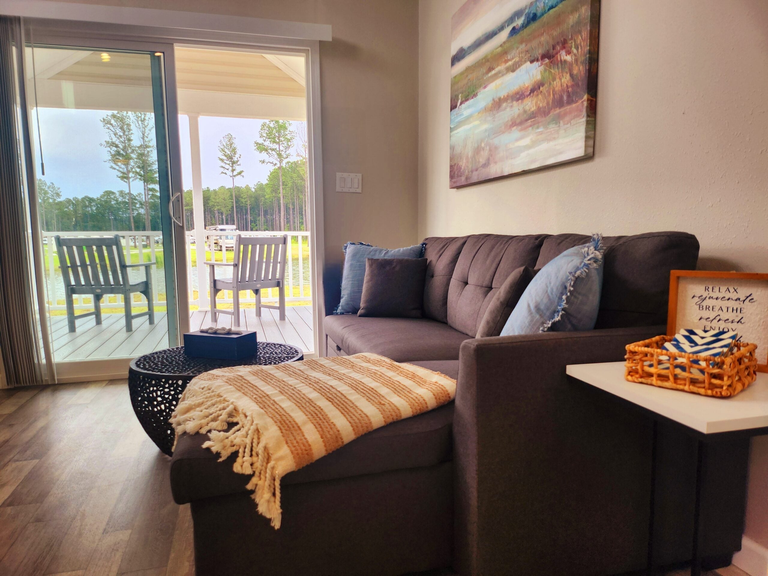 A cozy living room with a gray sectional sofa, a striped blanket, a round black coffee table, and a large window overlooking a porch with chairs and a view of trees.