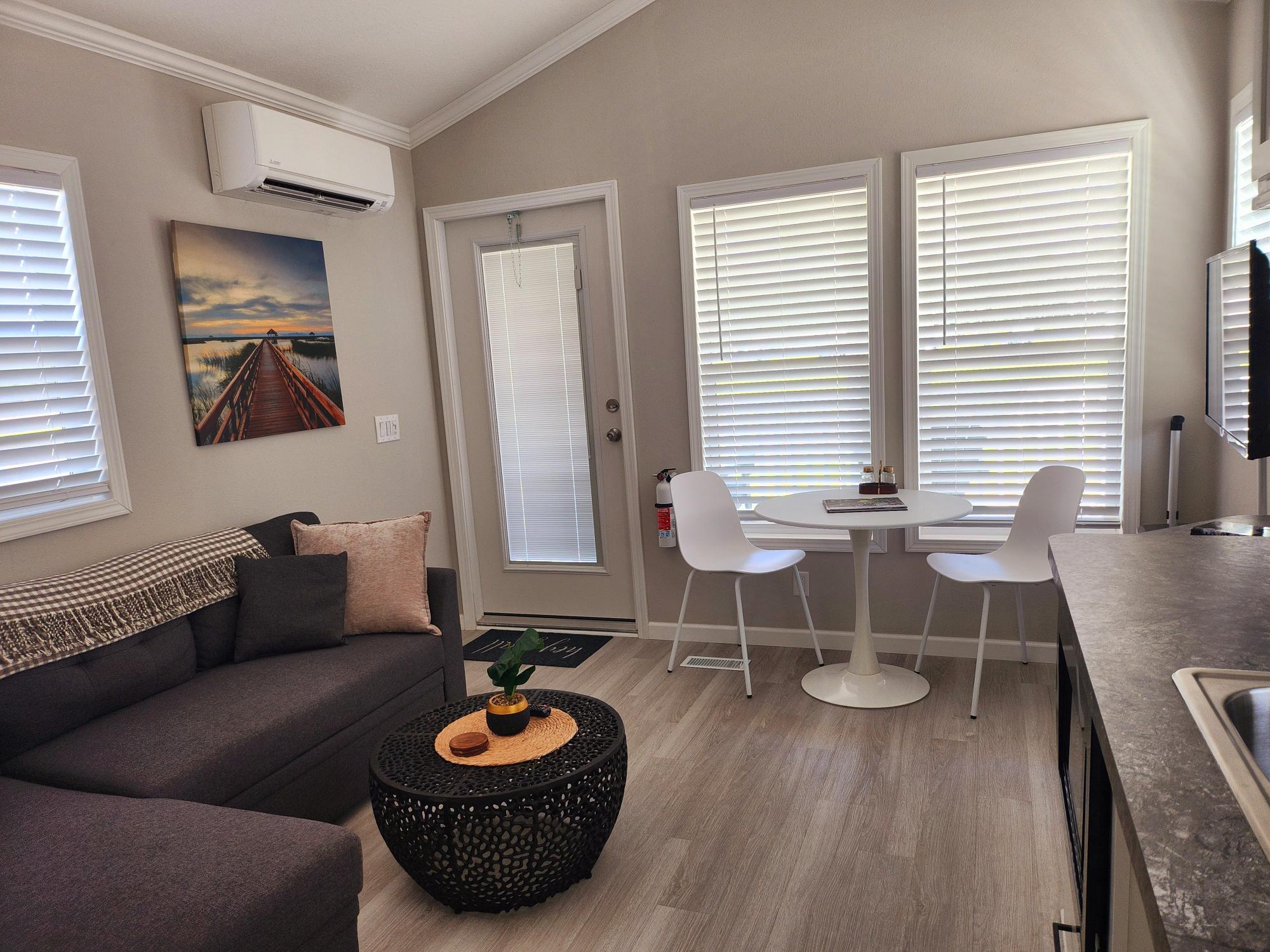 A small living room with a gray sectional sofa, round black coffee table, wall art, a white dining table with two chairs, and large windows with blinds.