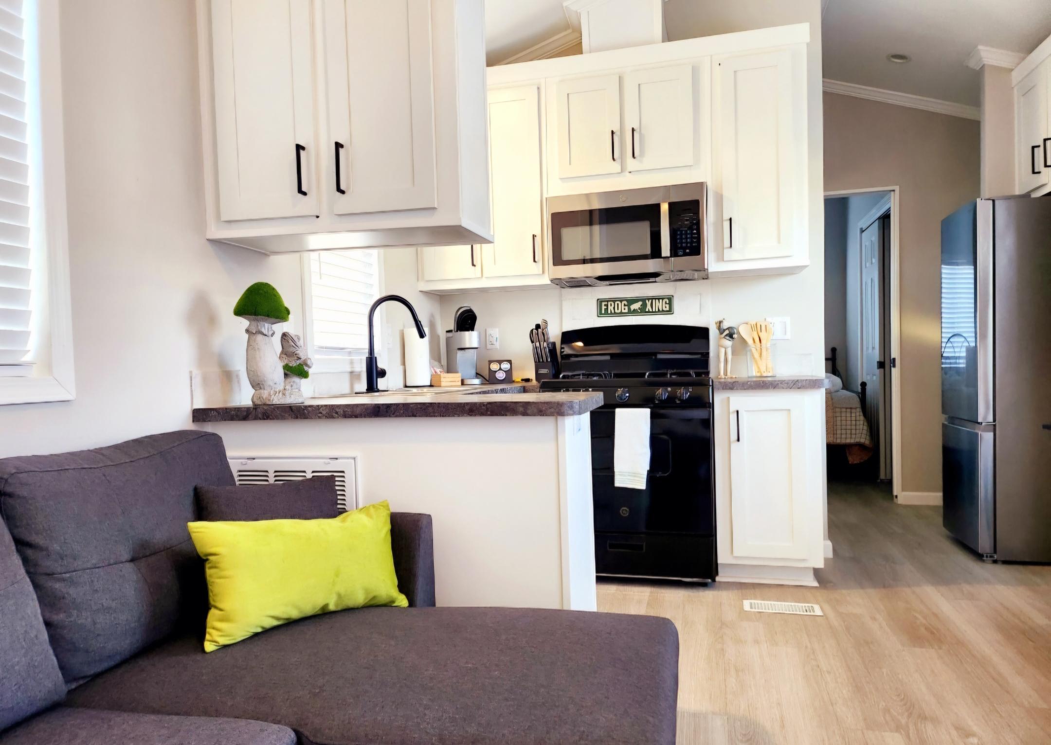 A compact kitchen with white cabinets, black appliances, and a brown countertop, next to a gray couch with a green pillow; a bedroom is visible through an open doorway.