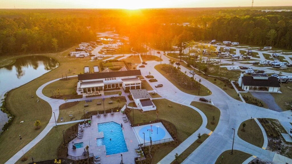 Aerial view of an RV park at sunset, featuring a swimming pool, modern buildings, winding roads, scattered RVs, and surrounding trees.