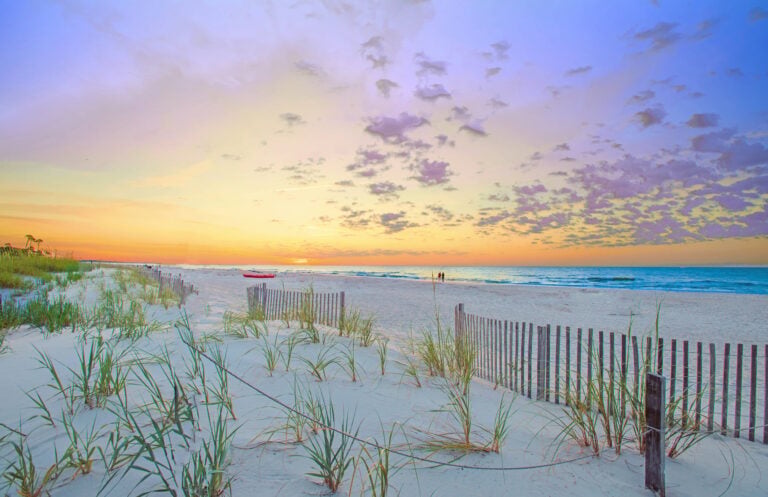 Sandy beach with dune grass, wooden fences, and a red boat at sunrise or sunset. Two people walk near the water under a colorful sky with scattered clouds.