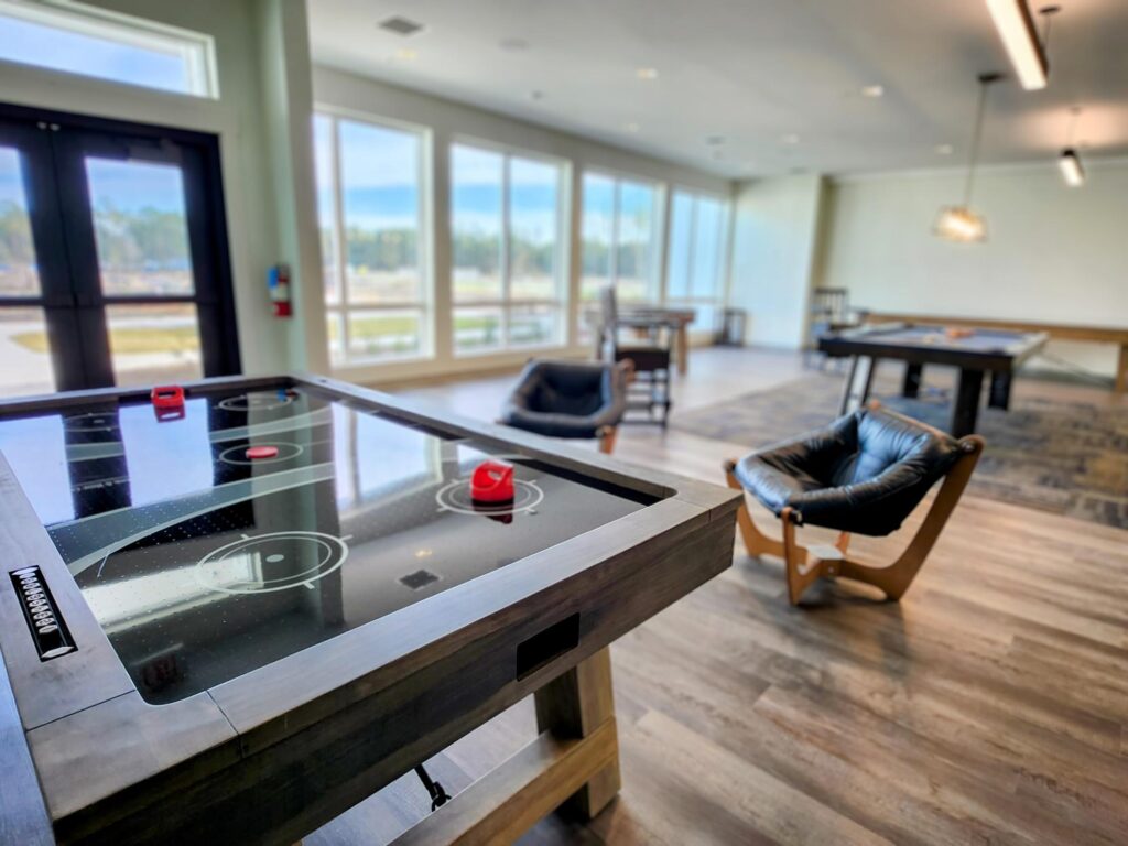 A modern game room with an air hockey table in the foreground, lounge chairs, large windows, and other game tables in the background.
