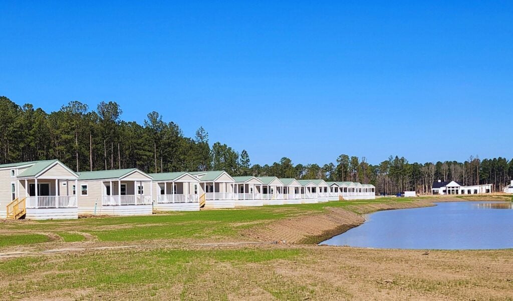 Row of small, light-colored modular homes with porches facing a pond, surrounded by grass and trees under a clear blue sky.