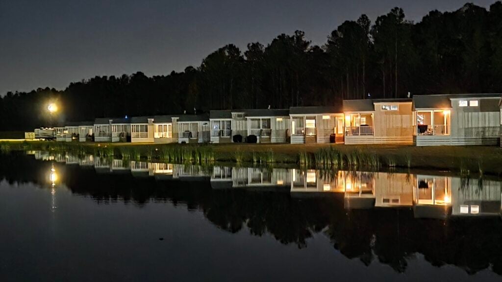 Row of illuminated lakeside cabins at night with their reflections visible in the calm water, trees silhouetted in the background.