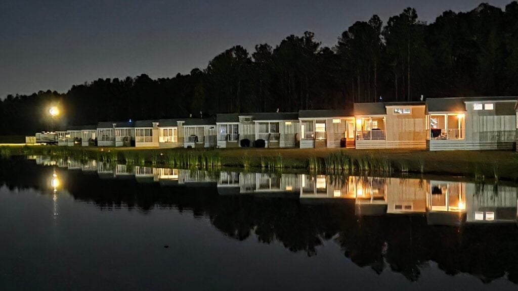 Row of modern lakeside homes at night with lights on, reflected in calm water; trees form a dark backdrop behind the houses.
