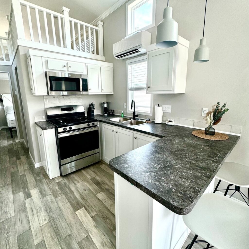 Modern kitchen with white cabinets, stainless steel appliances, dark countertops, pendant lights, and a breakfast bar with two stools. A loft area is visible above.