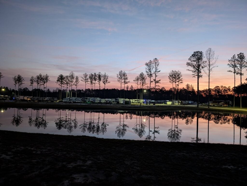 Sunset sky reflected in a calm lake, with a row of tall trees and RVs lined up in the background at a campground.