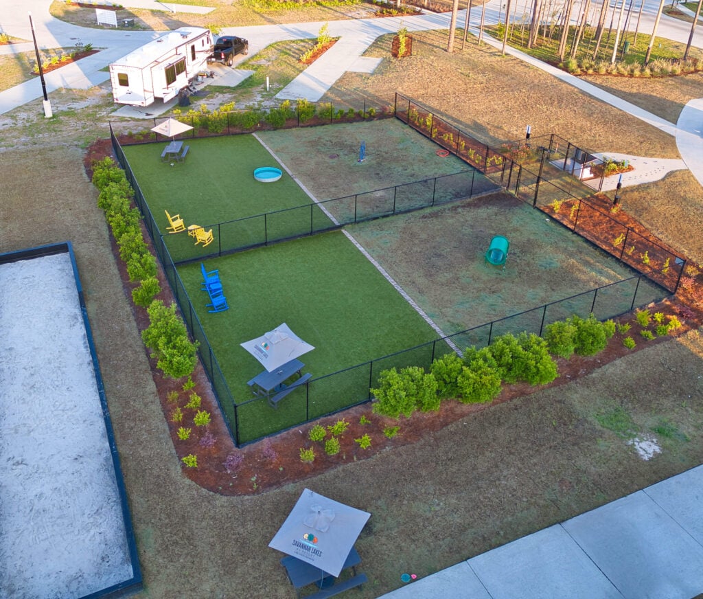 Aerial view of a fenced dog park with agility equipment, picnic tables, and benches. An RV and parking area are nearby. The surrounding area has grass, shrubs, and paved walkways.
