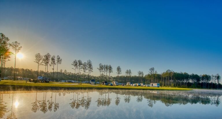 RVs and vehicles are parked along a tree-lined shore at sunrise, reflected in the calm water of a lake under a clear blue sky.