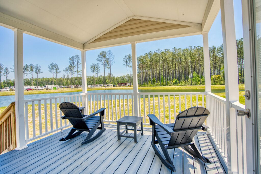Two black Adirondack chairs and a small table on a covered porch overlook a pond and trees on a sunny day.