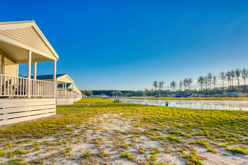 Single-story cabins with covered porches face a calm lake, with grassy ground in the foreground and parked RVs across the water under a clear blue sky.