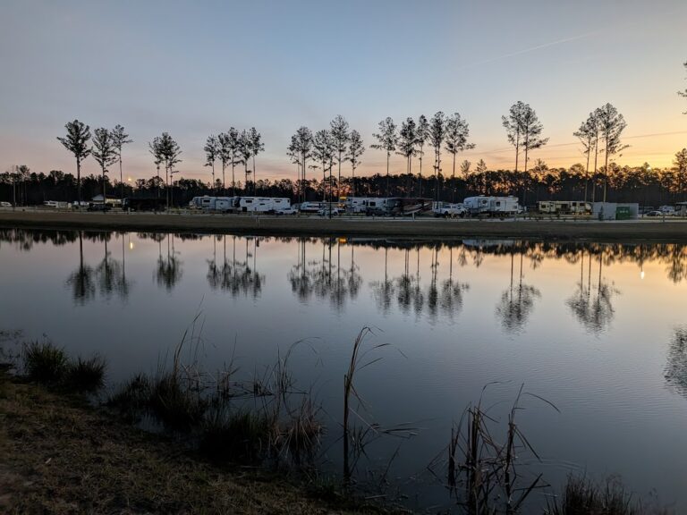 A row of RVs is parked near a pond at sunset, with tall trees and their reflections visible in the calm water.