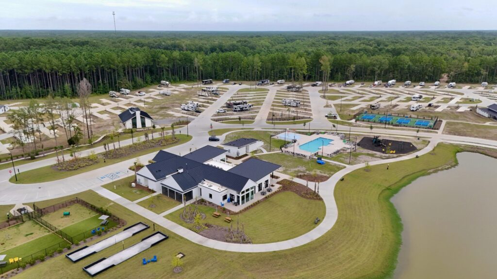 Aerial view of an RV park with parked RVs, a main building, a pool, landscaped green areas, and a pond, surrounded by a forest.