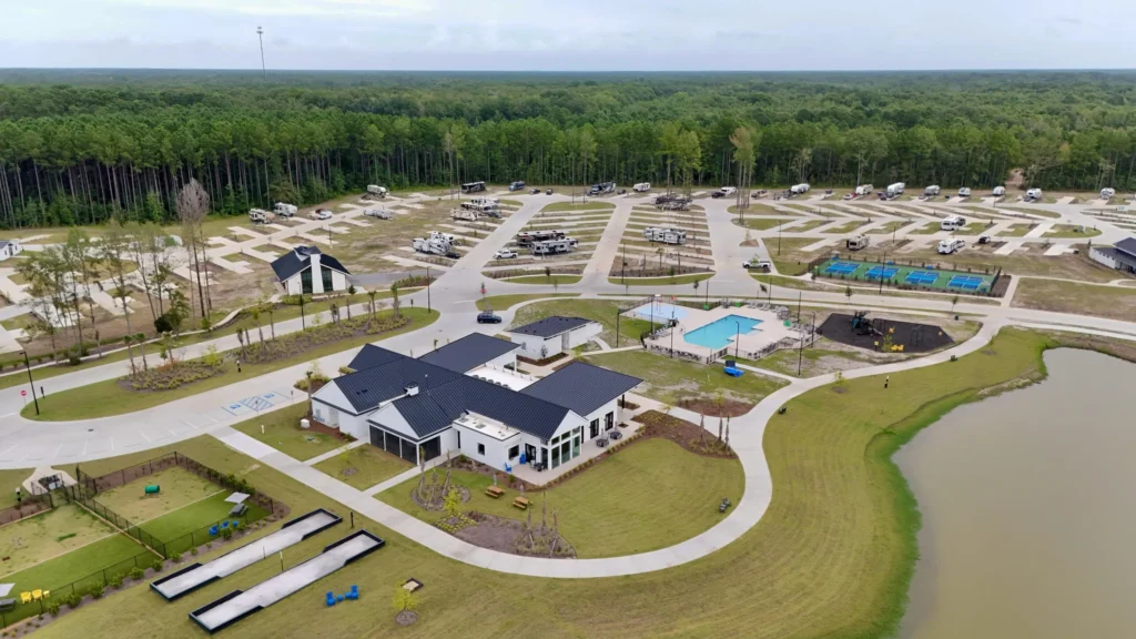 Aerial view of an RV park with a central building, outdoor pool, parking spaces, scattered RVs, and surrounding green areas near a wooded forest.