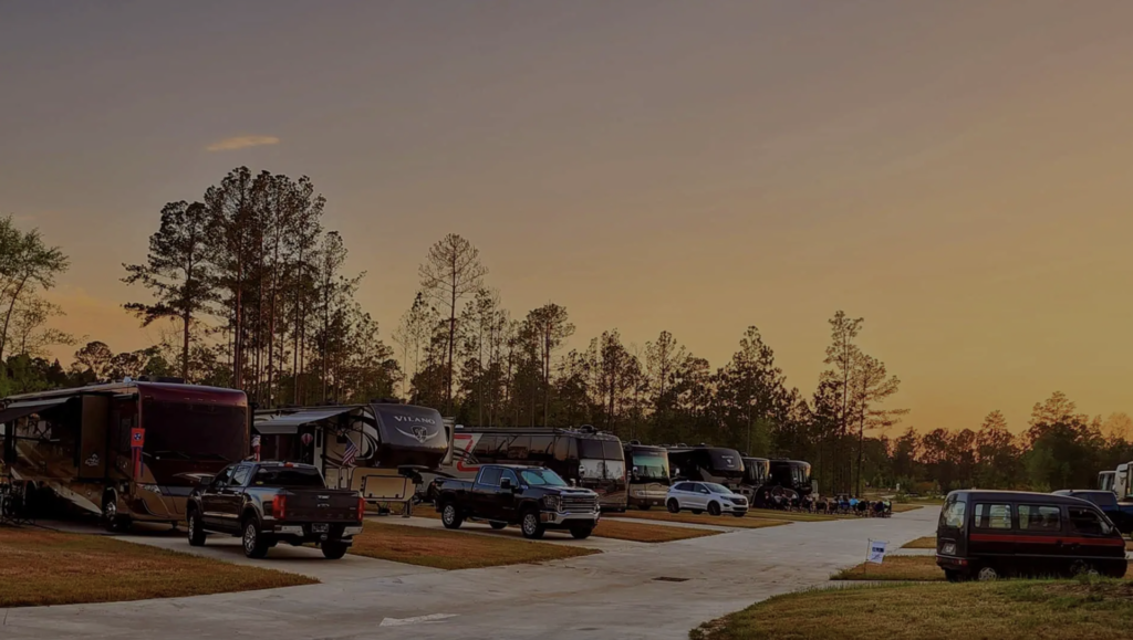 Row of RVs and trucks parked along a paved road at a campground, with trees and a clear sky at sunset in the background.