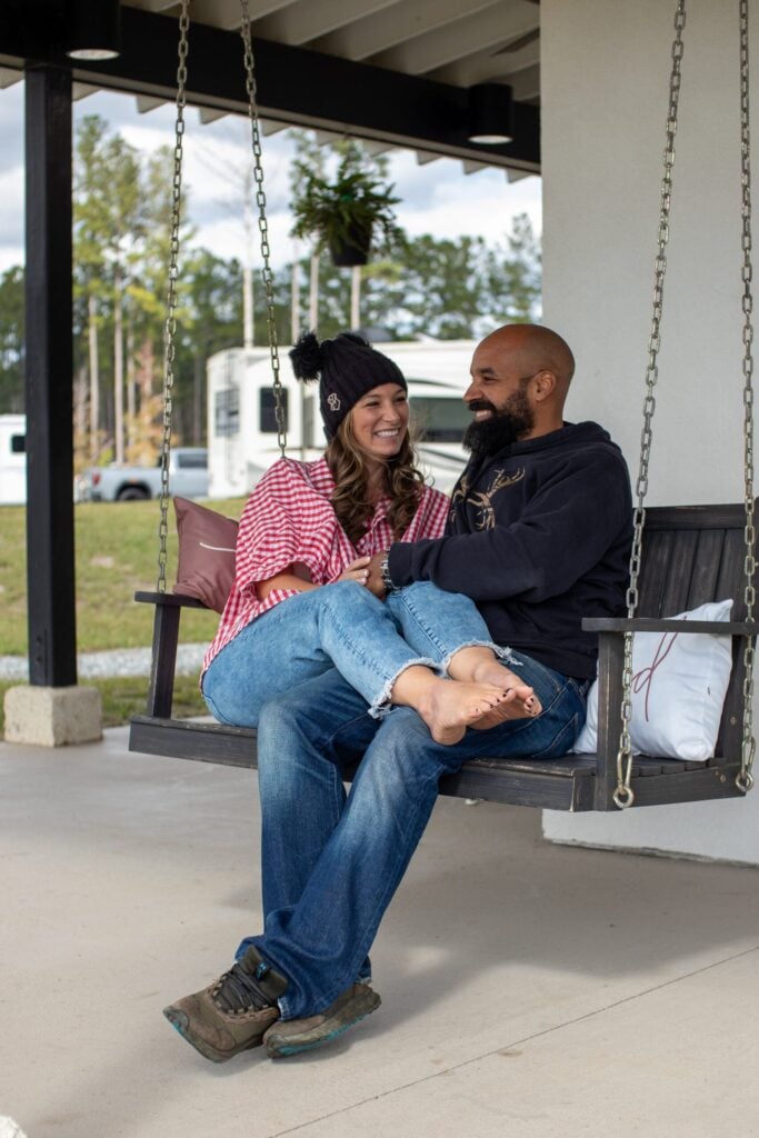 A woman and man sit together on a porch swing, smiling and facing each other. The woman is barefoot and wearing a beanie, while the man wears jeans and boots.
