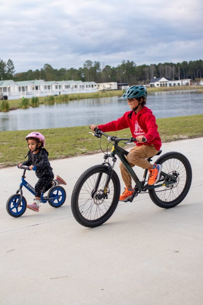 A young boy on a bicycle and a young girl on a balance bike ride on a paved path near a lake, both wearing helmets.