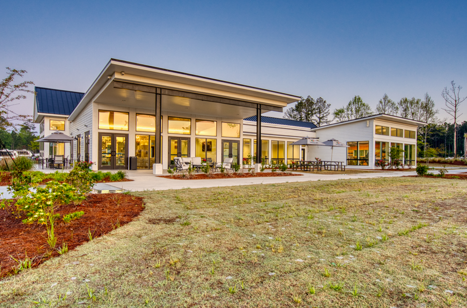 Modern building with large windows and outdoor seating, surrounded by landscaping and a grassy area, photographed at dusk.
