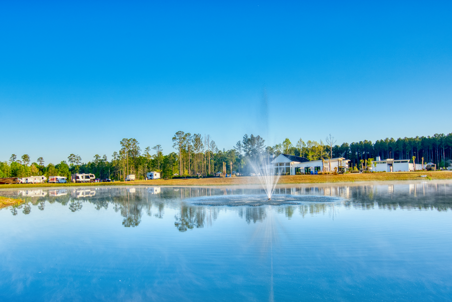 A pond with a water fountain in the center, reflecting nearby RVs, trees, and buildings under a clear blue sky.