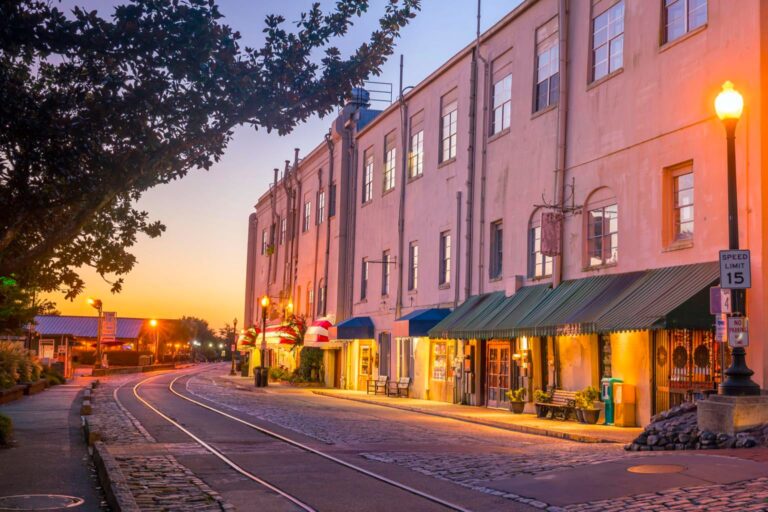 A cobblestone street with tram tracks runs alongside historic buildings at sunset, illuminated by streetlights and storefront lights.
