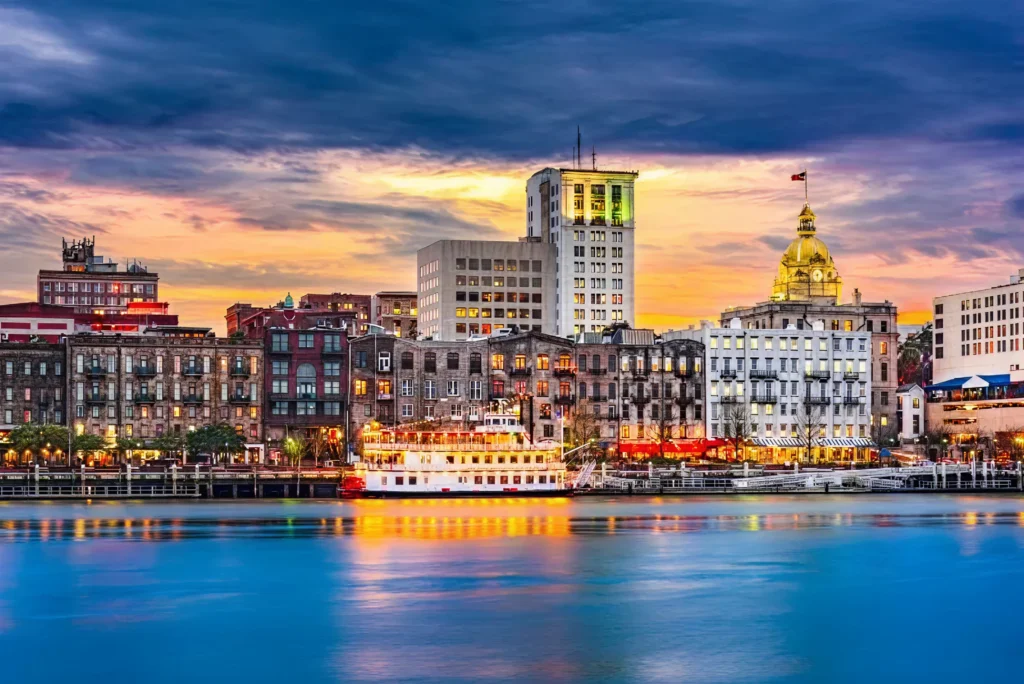 Savannah, Georgia waterfront at sunset, featuring historic buildings, a riverboat on the water, and the golden dome of Savannah City Hall under a dramatic, cloudy sky.