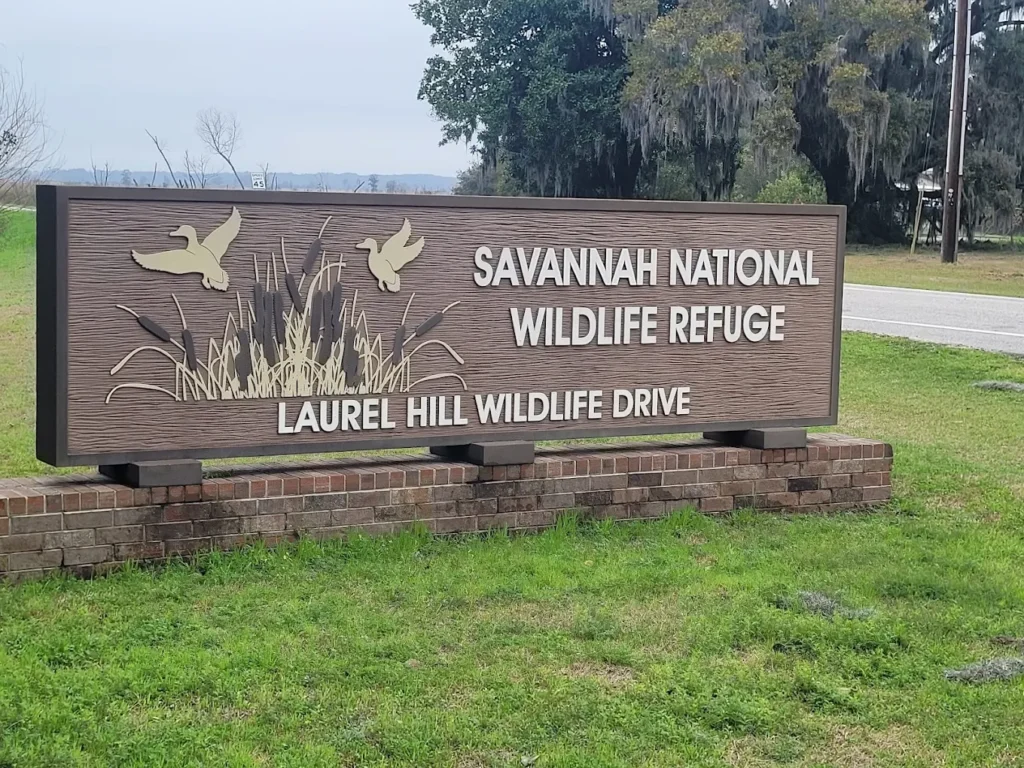 A wooden sign reads "Savannah National Wildlife Refuge, Laurel Hill Wildlife Drive" with images of flying birds and grass, situated on a grassy area near a road.