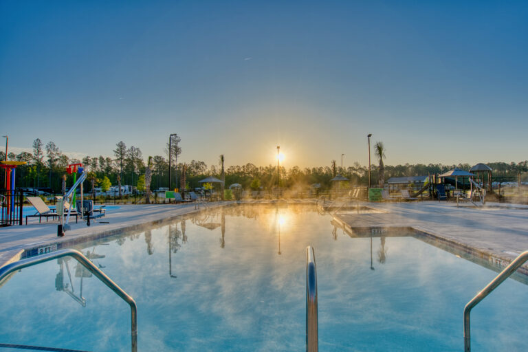 Outdoor swimming pool with handrails and steam rising, surrounded by trees and playground equipment, at sunrise under a clear sky.