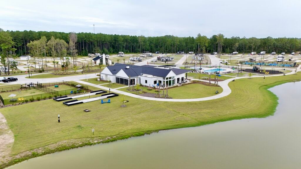 A modern clubhouse with a pool and playground sits beside a pond, surrounded by green lawns, walking paths, and parked RVs in a campground setting.