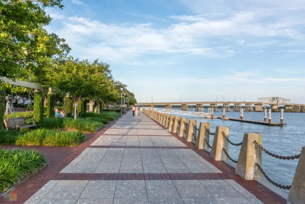 A paved waterfront walkway lined with trees, benches, and posts with chains, runs alongside a river under a partly cloudy sky.