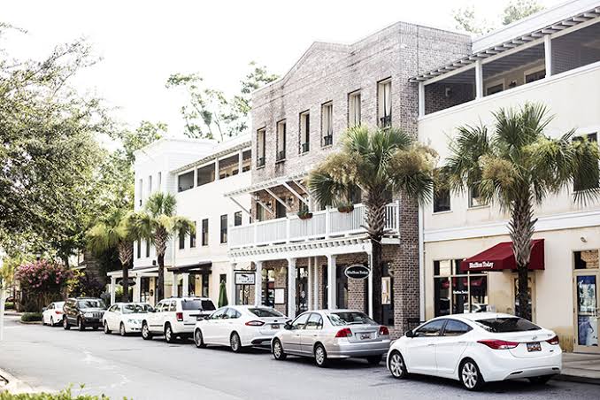 A row of parked cars lines the street in front of a mixed-use building with shops on the ground floor and palm trees along the sidewalk.