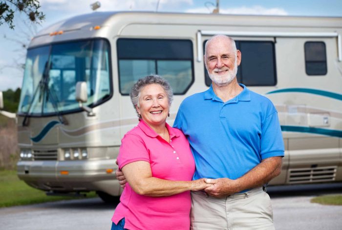 An elderly couple stands in front of a large RV, smiling and holding hands. The man is wearing a blue polo shirt and beige pants, and the woman is wearing a pink polo shirt and beige pants. Snowbirds enjoying their adventures, they bask in the sunny day with a partly cloudy sky.